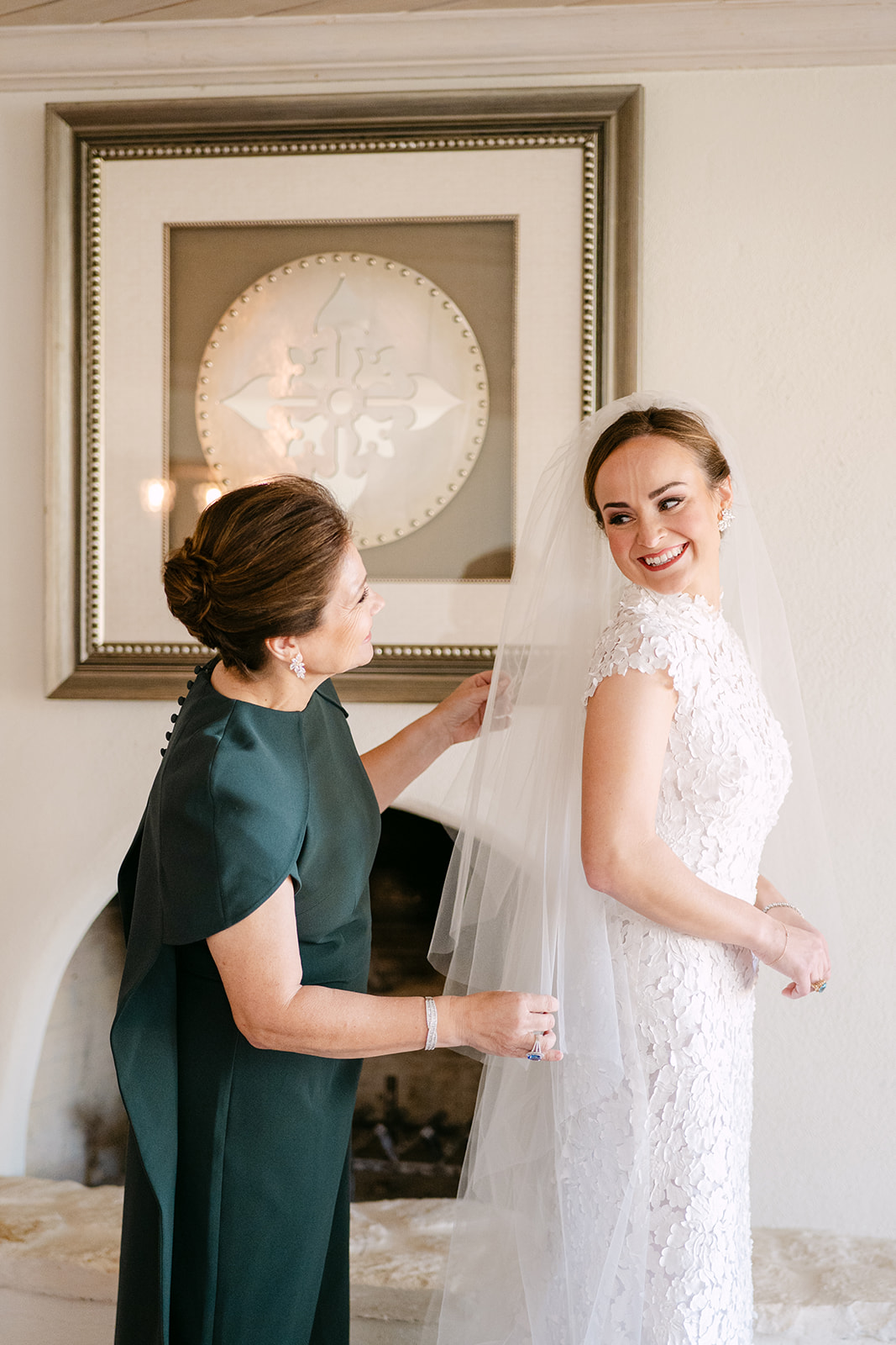 A woman in a dark dress helps a bride in a lace gown with her dress. The bride smiles at the camera, standing in a room with a framed decorative plate on the wall.