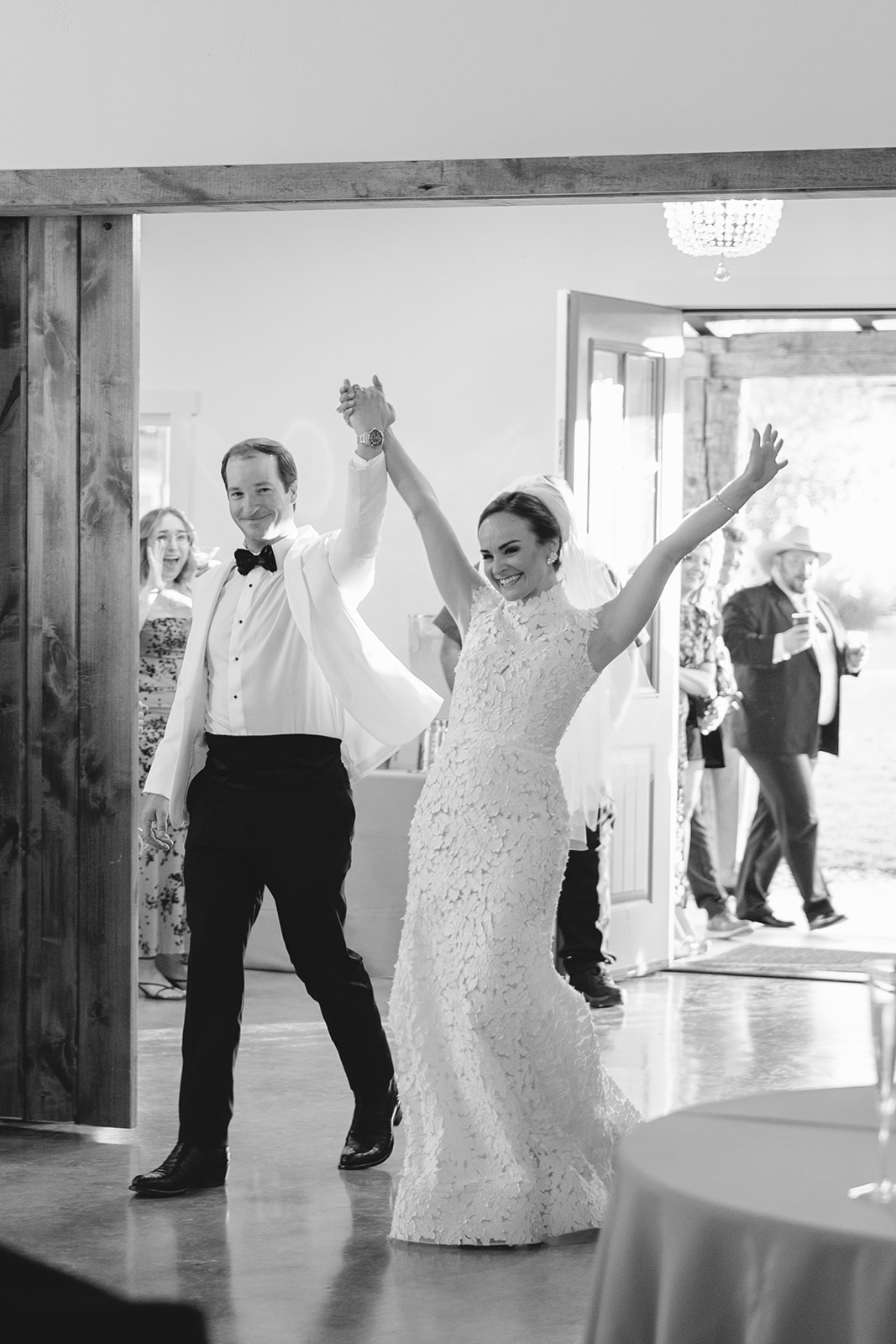 A bride and groom enter a room through wooden doors, smiling and waving. A tiered cake and floral arrangements are visible in the foreground.