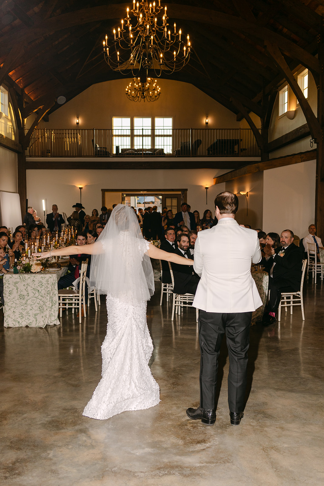 A bride and groom enter a room through wooden doors, smiling and waving. A tiered cake and floral arrangements are visible in the foreground.