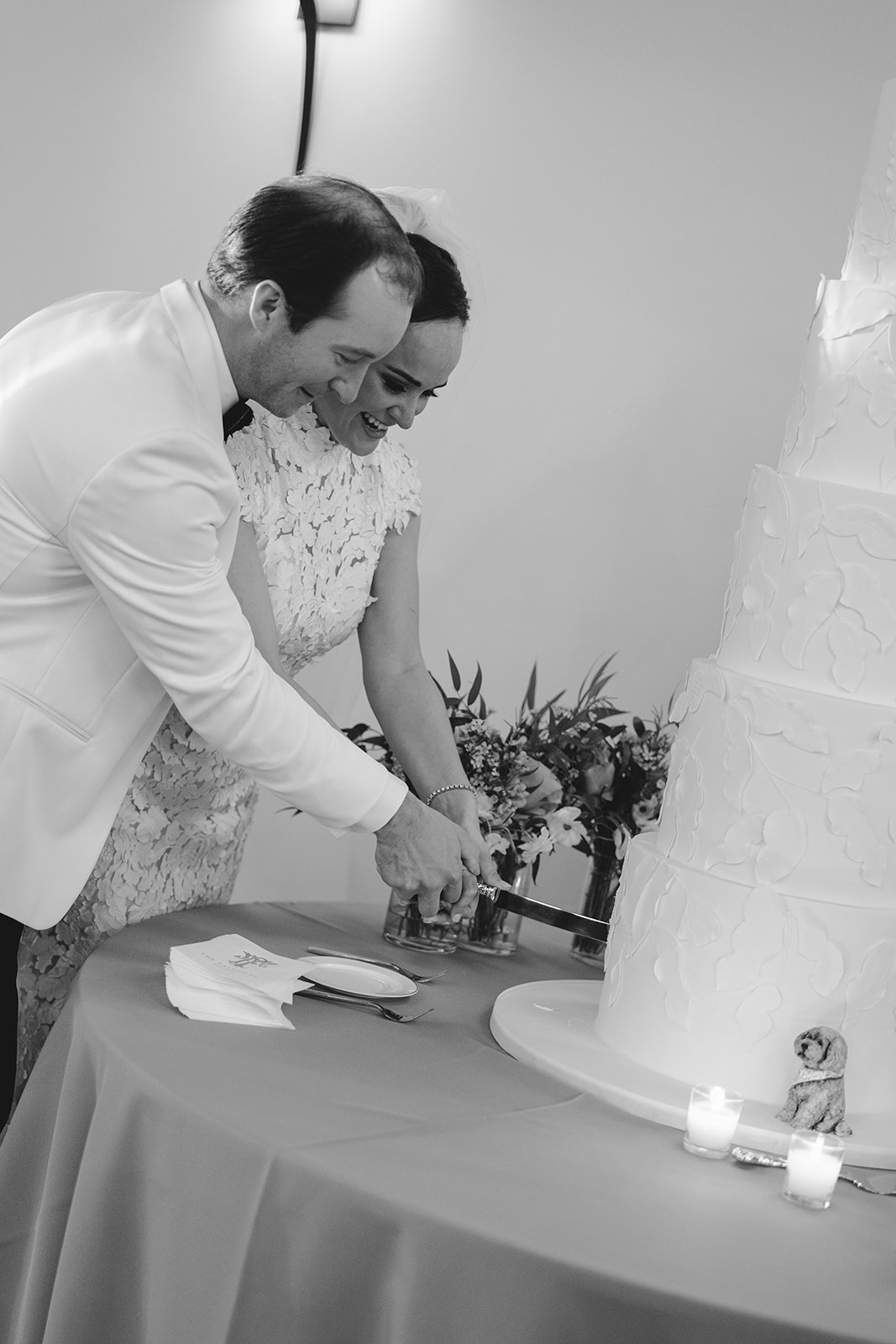 A couple in formal attire cuts a tall, multi-tiered wedding cake on a table with flowers.