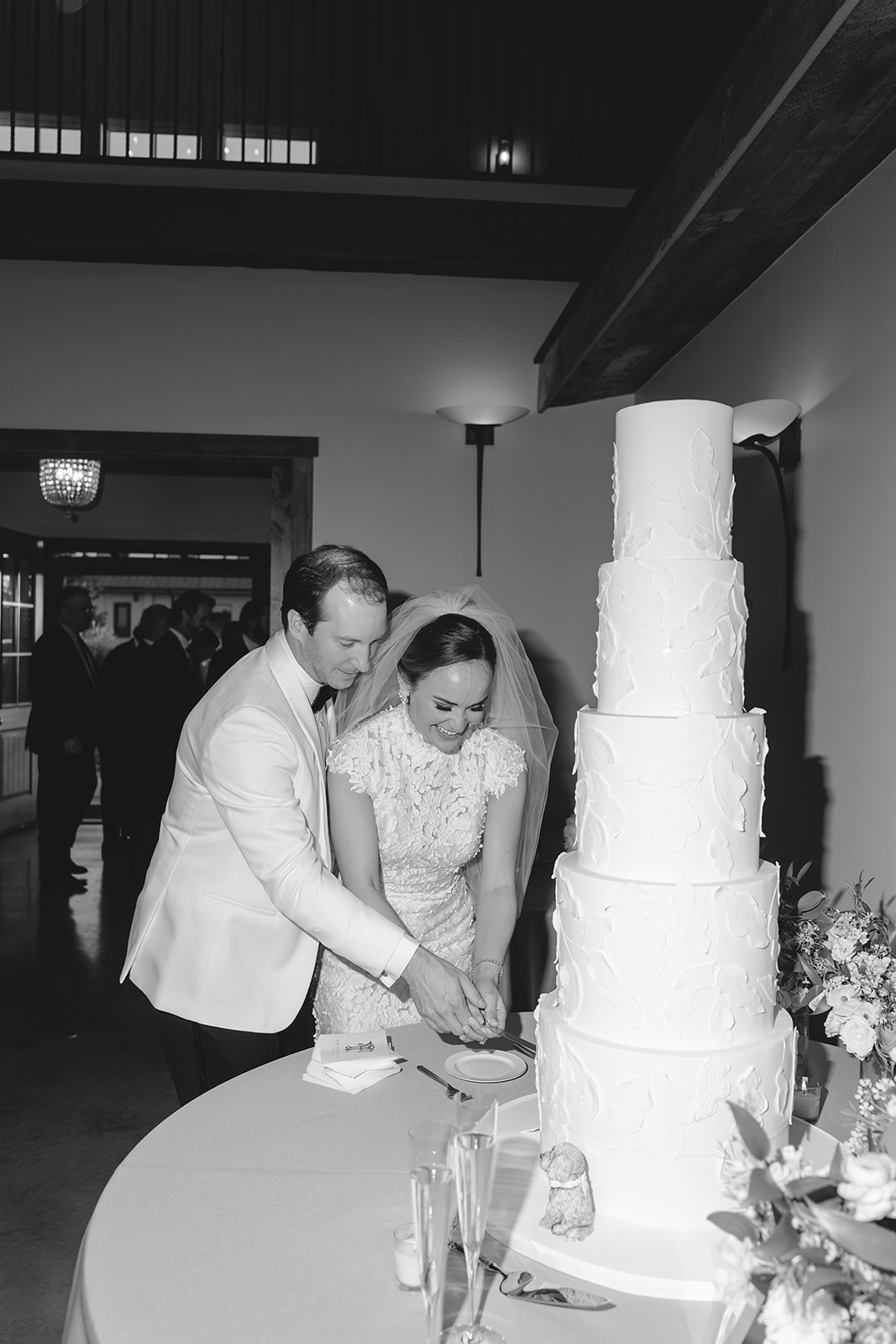 A couple in formal attire cuts a tall, multi-tiered wedding cake on a table with flowers.