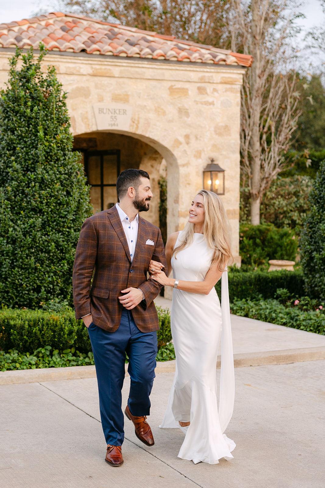 A man in a brown jacket and blue pants stands next to a woman in a white dress in front of a stone building with greenery.