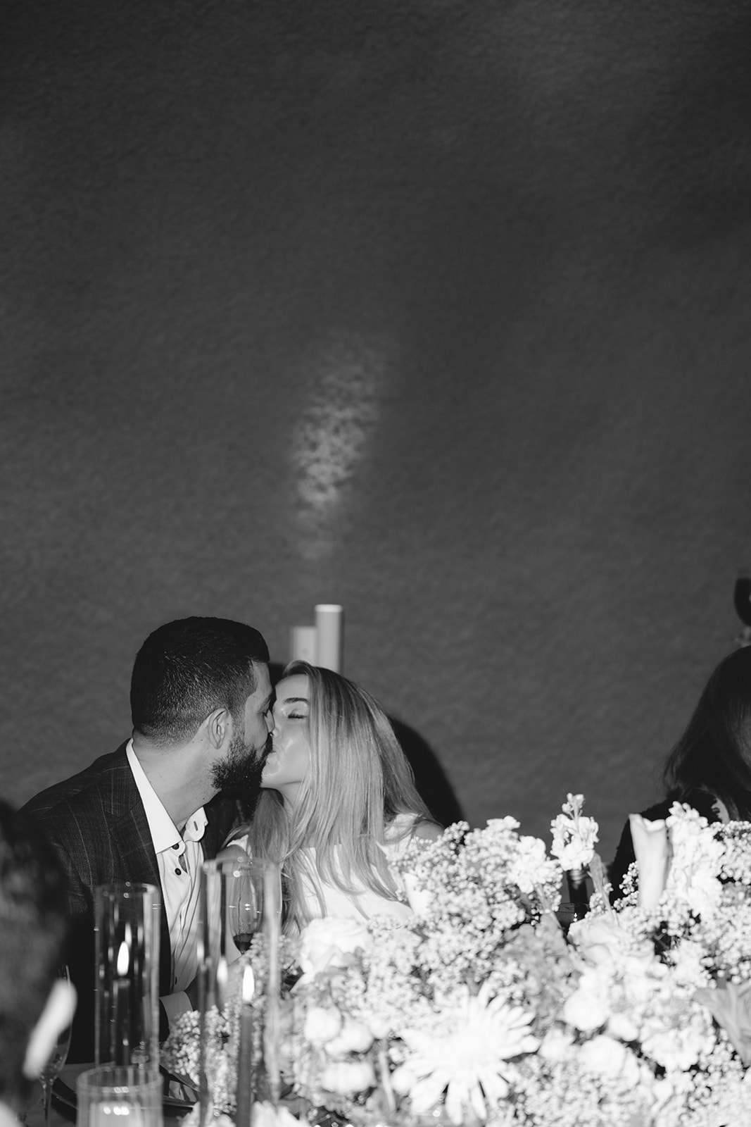 A couple shares a kiss at a table adorned with flowers and candles in a black and white setting.