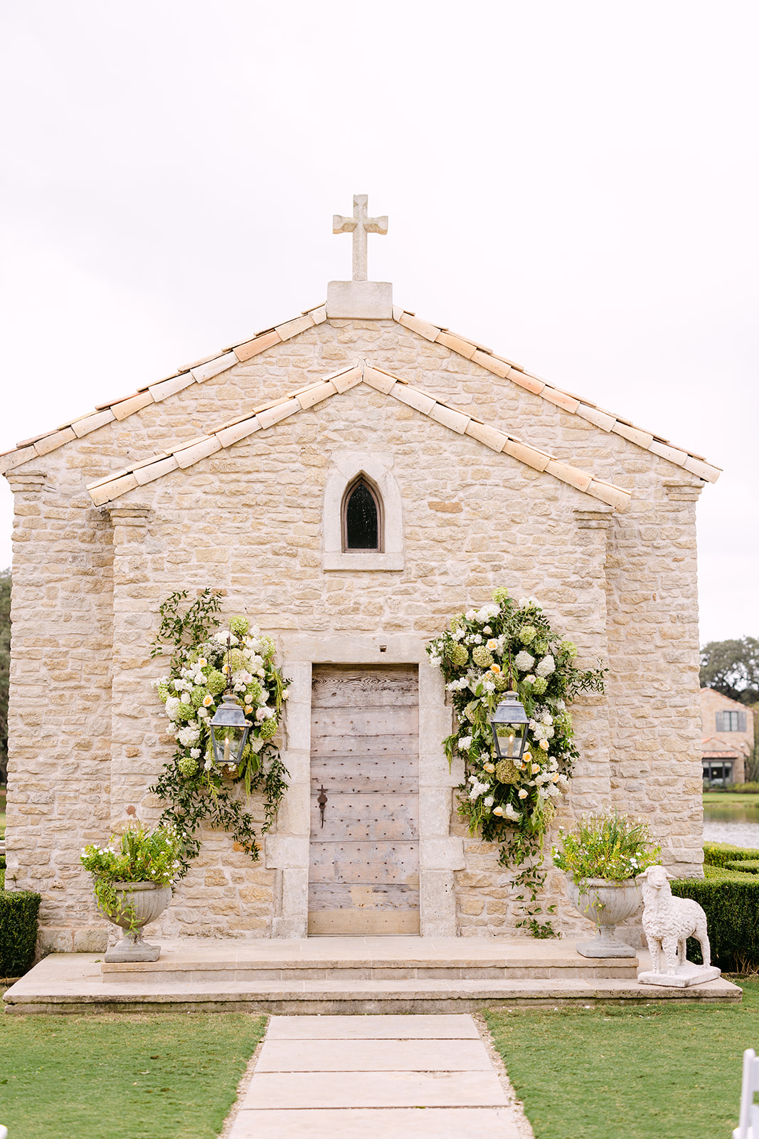 A small stone chapel with a cross on top. The entrance is adorned with green and white floral arrangements. Two planters and a stone lion statue are on either side of the path leading to the door.