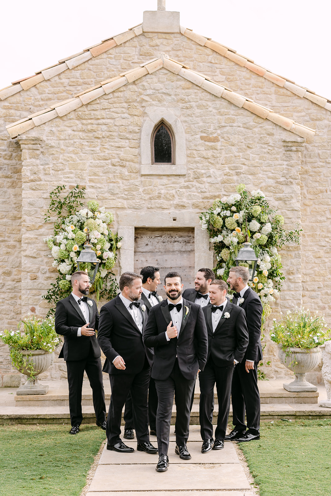 Seven groomsmen in black tuxedos stand in front of a stone chapel with floral arrangements.