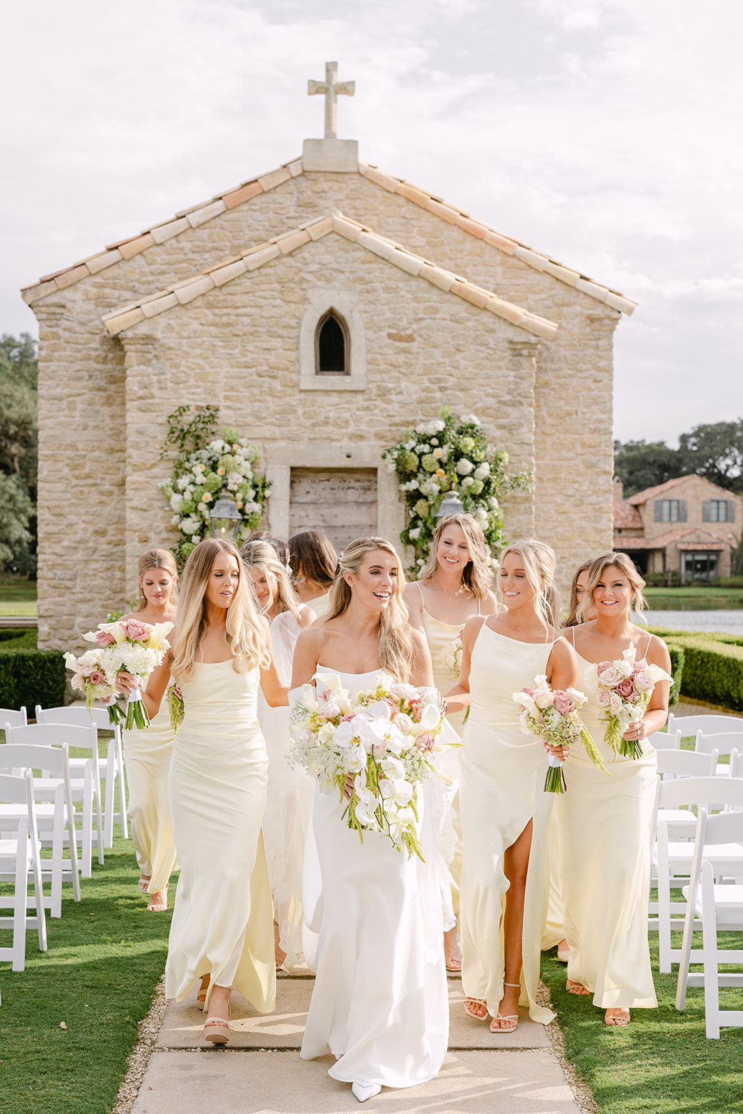 A wedding party stands in front of a stone chapel. The bridesmaids wear light yellow dresses, while the groomsmen and other men are in black suits. The couple is in the center.