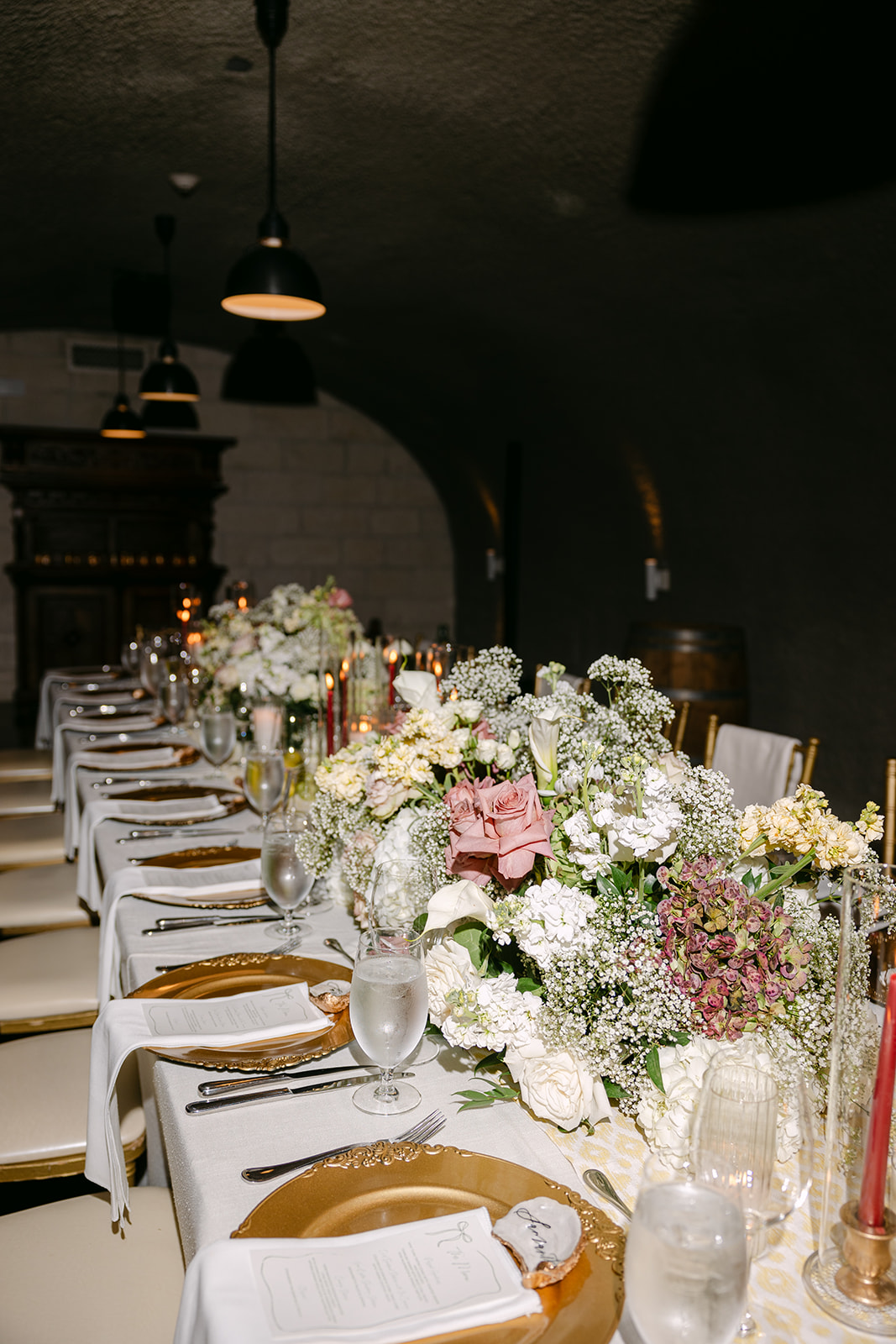Elegant dining table set in a dimly lit room with curved arches, adorned with floral centerpieces, red candles, and elegant tableware.