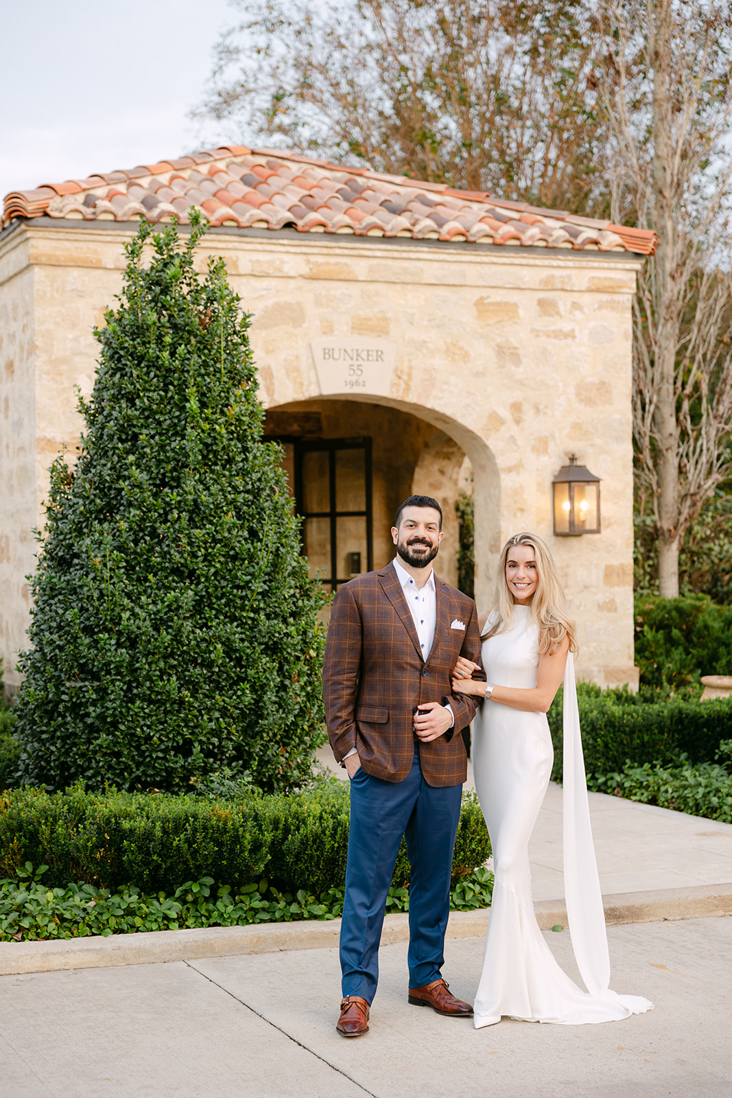 A man in a brown jacket and blue pants stands next to a woman in a white dress in front of a stone building with greenery.