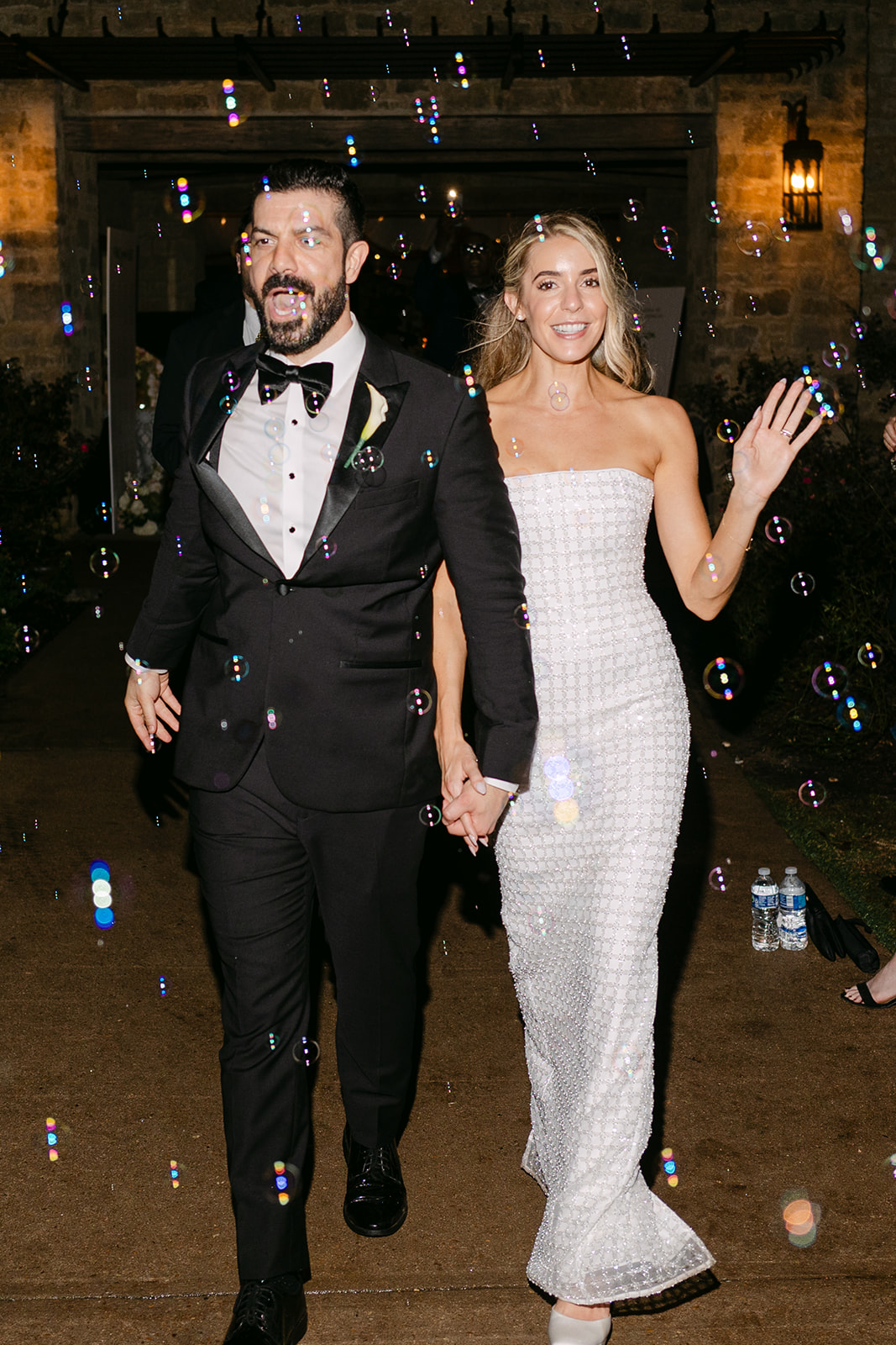 A newlywed couple in formal attire walks hand in hand through guests blowing bubbles at an outdoor event.