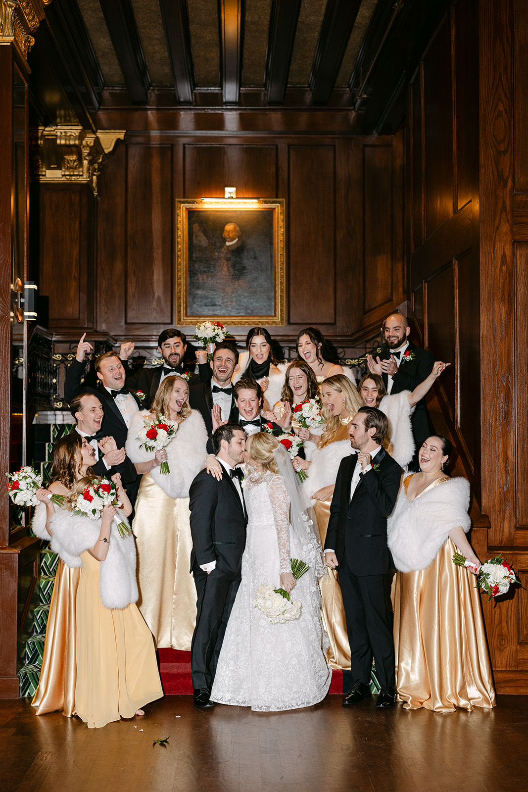 A wedding party poses on a staircase in formal attire; the bride and groom stand at the center, flanked by bridesmaids in gold dresses and groomsmen in black suits, all holding bouquets.