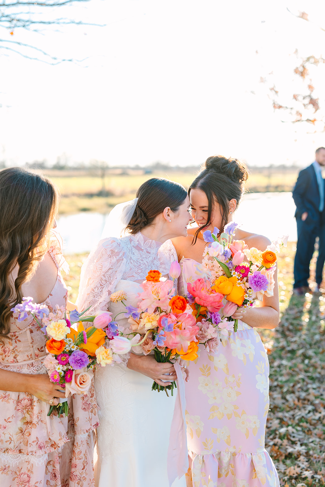 Three women in floral dresses stand outdoors, smiling and holding colorful bouquets of flowers, with a pond and field in the background for a wedding at Beckendorff Farms in Katy, TX