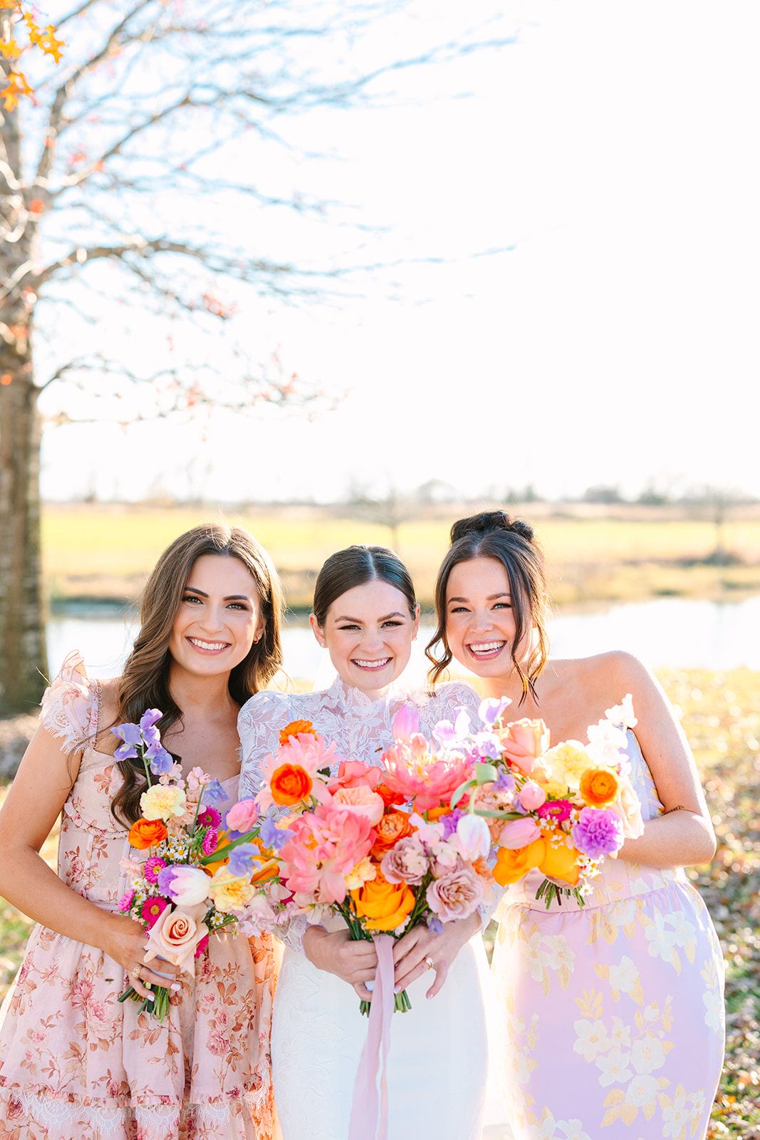 Three women in floral dresses stand outdoors, smiling and holding colorful bouquets of flowers, with a pond and field in the background for a wedding at Beckendorff Farms in Katy, TX