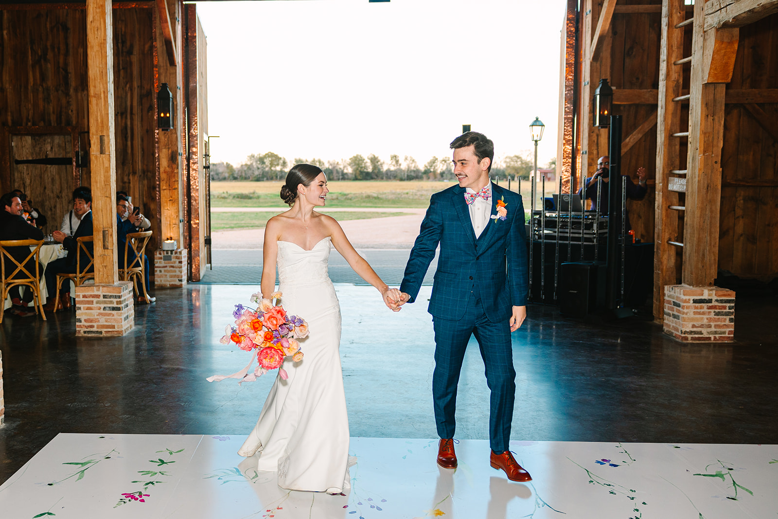 A bride in a white dress and a groom in a blue suit hold hands and enter a barn venue with open doors, smiling at each other.