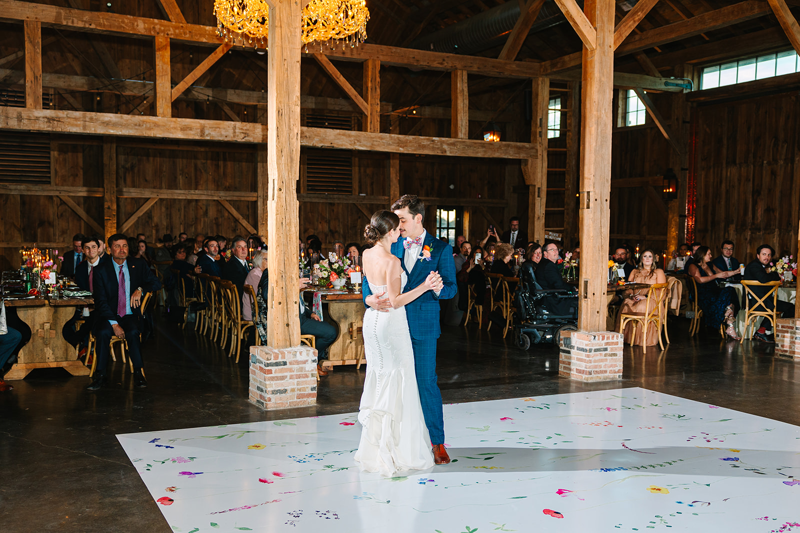 A bride and groom share their first dance on a decorated white floor in a rustic barn venue, with seated guests watching.