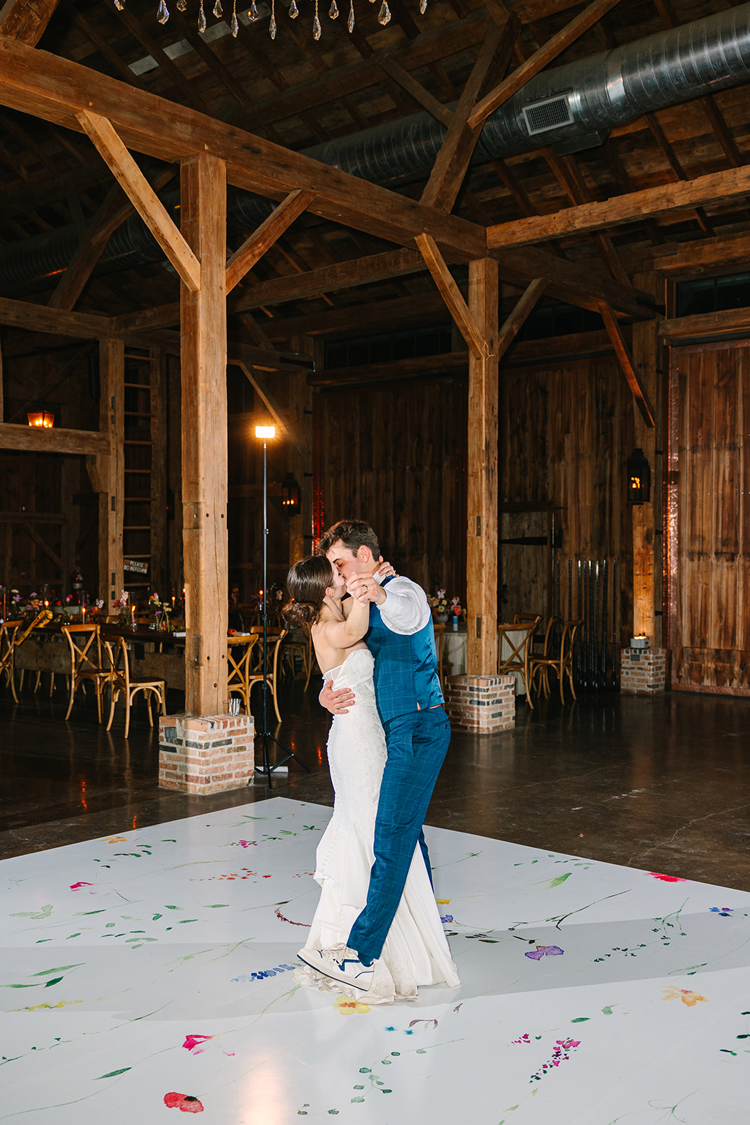 A bride and groom share a first dance on a white floor scattered with confetti in a rustic, wooden venue.