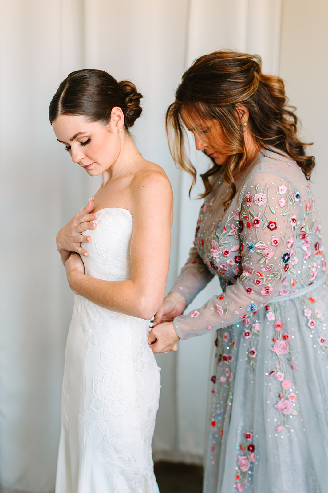 A woman in a gray floral dress helps a bride in a strapless white gown fasten the back of her dress in a softly lit room.