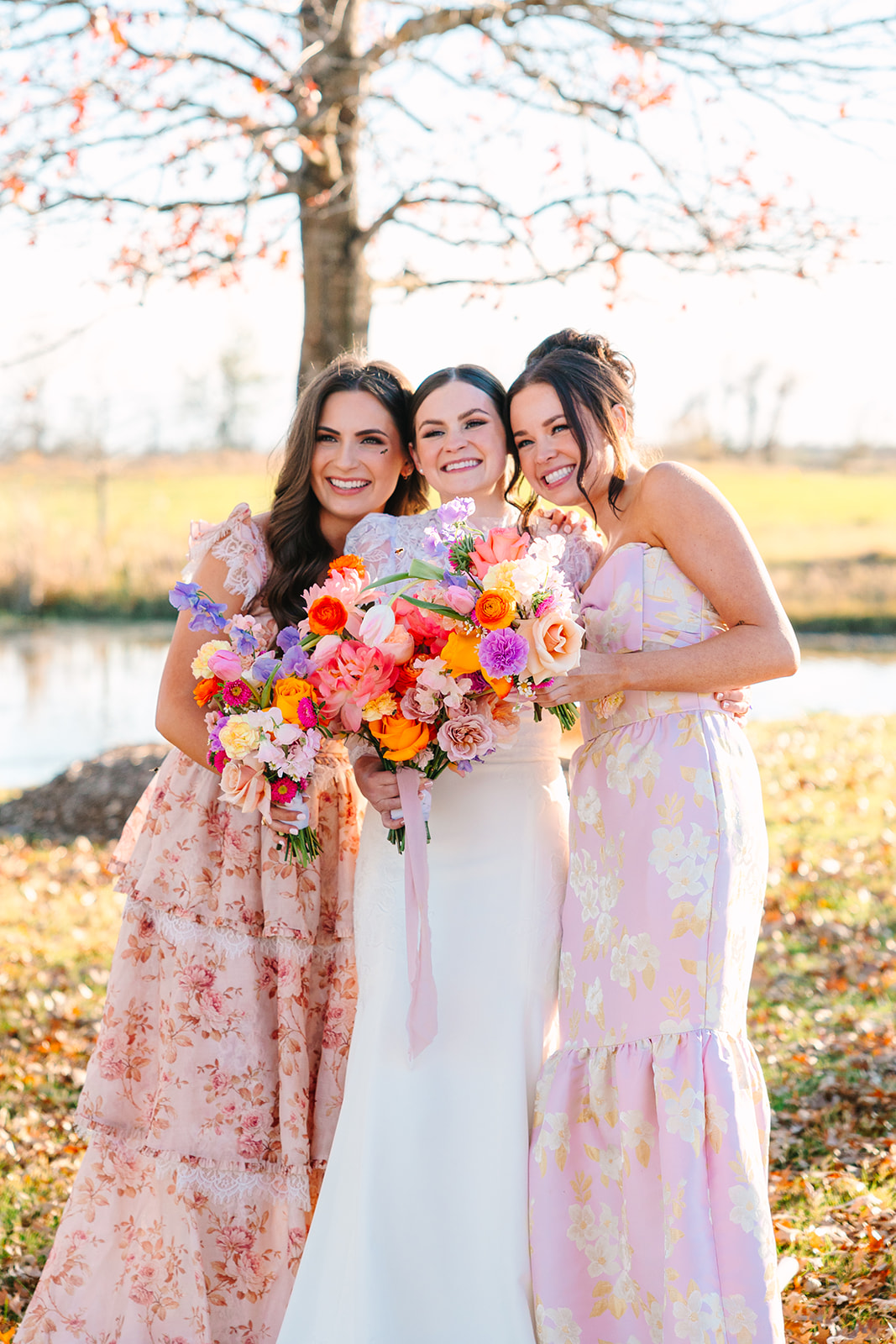 Three women in floral dresses stand outdoors, smiling and holding colorful bouquets of flowers, with a pond and field in the background for a wedding at Beckendorff Farms in Katy, TX