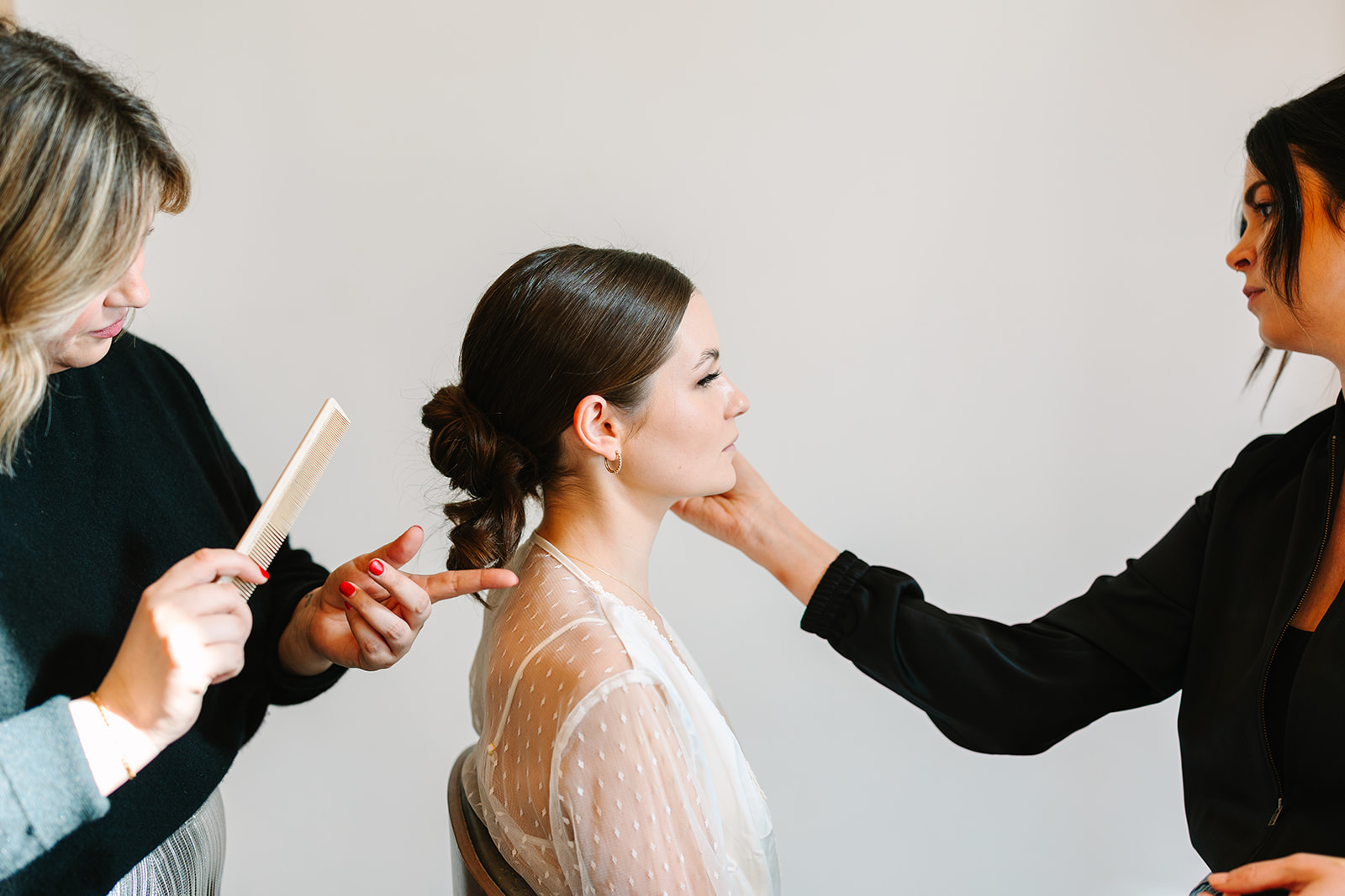A makeup artist applies false eyelashes to a woman wearing a white blouse, seated indoors with soft lighting in the background.