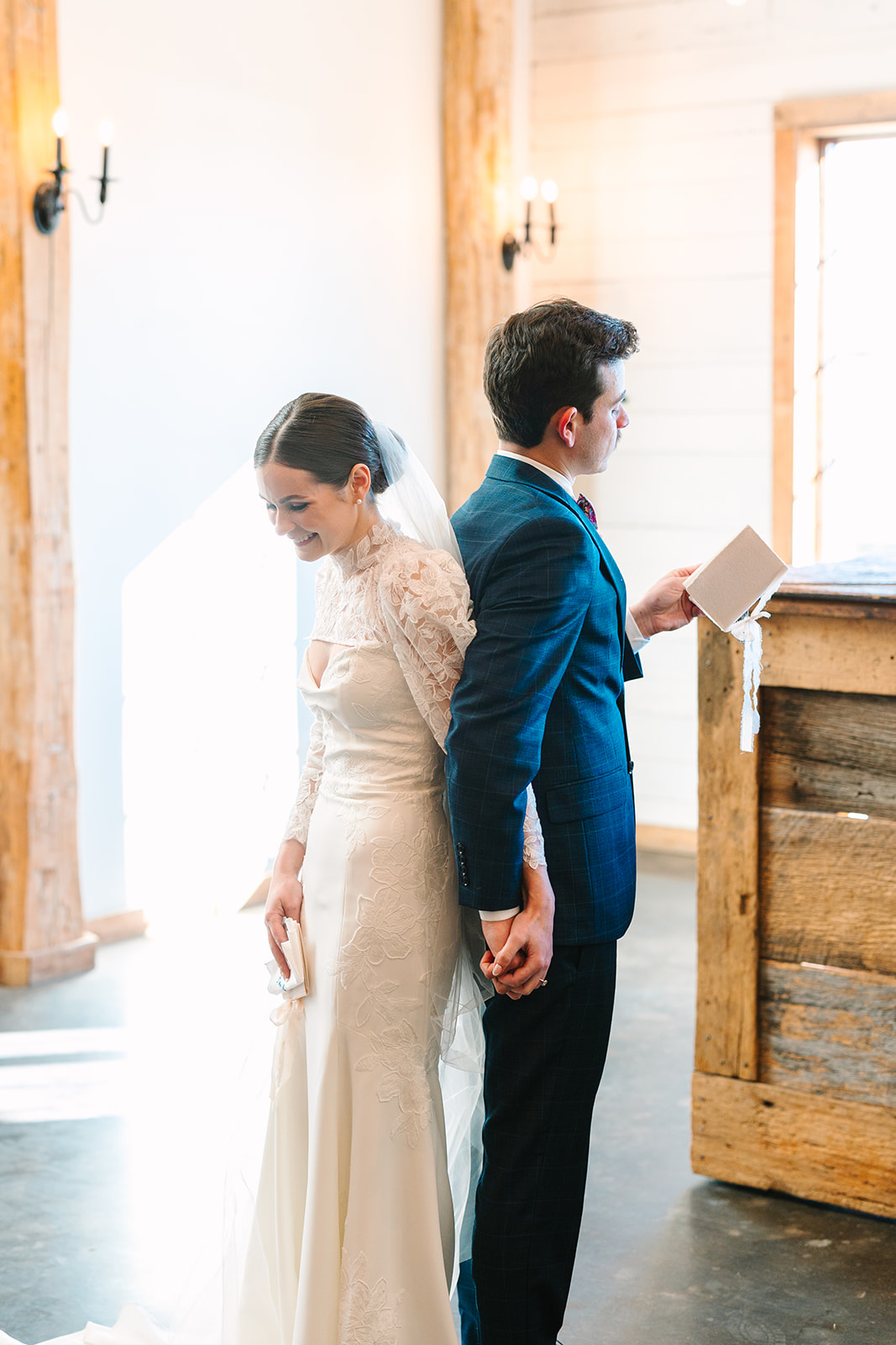 A bride and groom stand back-to-back indoors, dressed in wedding attire, with natural light illuminating the scene.
