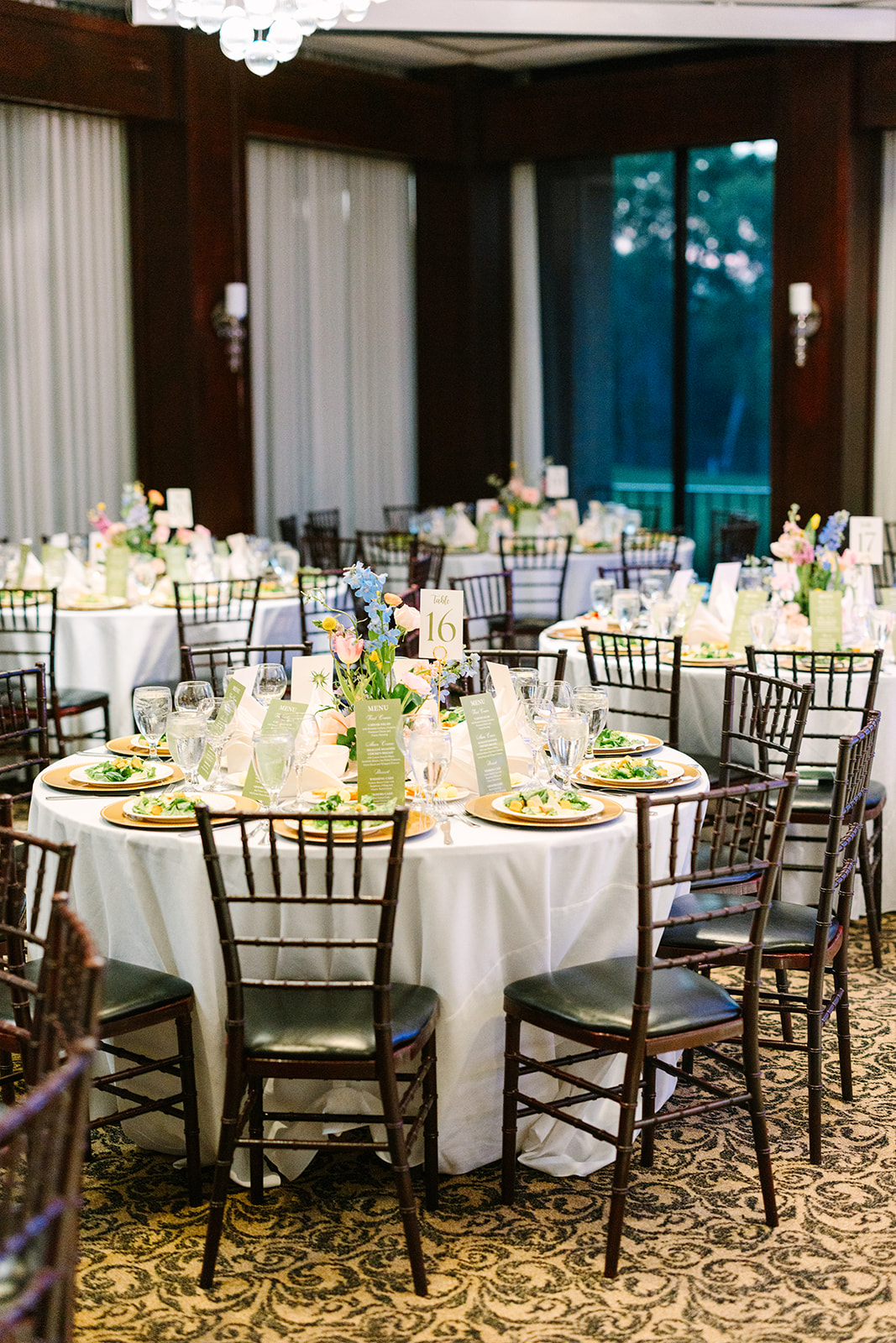 Round tables with white tablecloths are set for a formal event, featuring place settings, glassware, floral centerpieces, and dark wooden chairs in an elegant indoor venue at Elegant Braeburn Country Club Houston Wedding