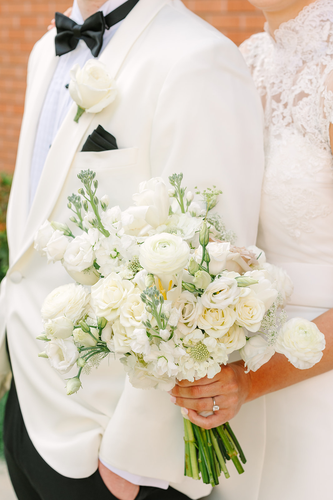 A bride in a white dress and veil stands next to a groom in a white tuxedo jacket, both smiling and posing outdoors in front of a brick building for their Braeburn Country Club Houston Wedding