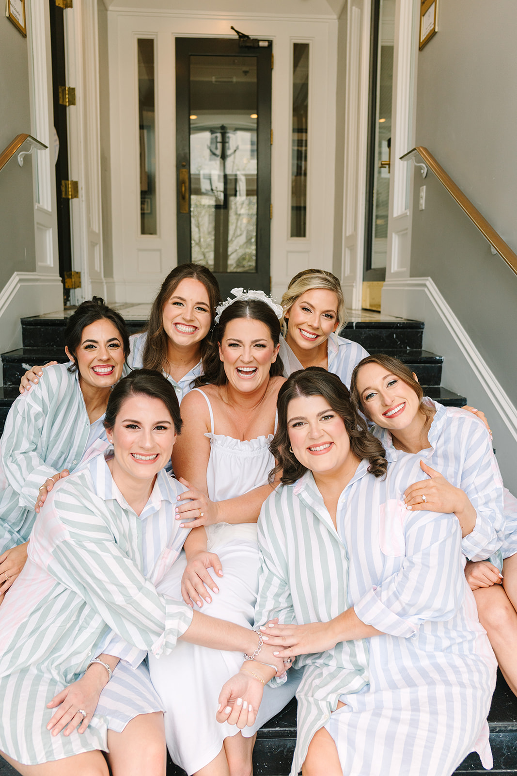 A bride in a white dress sits on steps with six bridesmaids wearing matching striped shirts and white socks, all posing and smiling for a group photo indoors for a wedding at the tremont hotel
