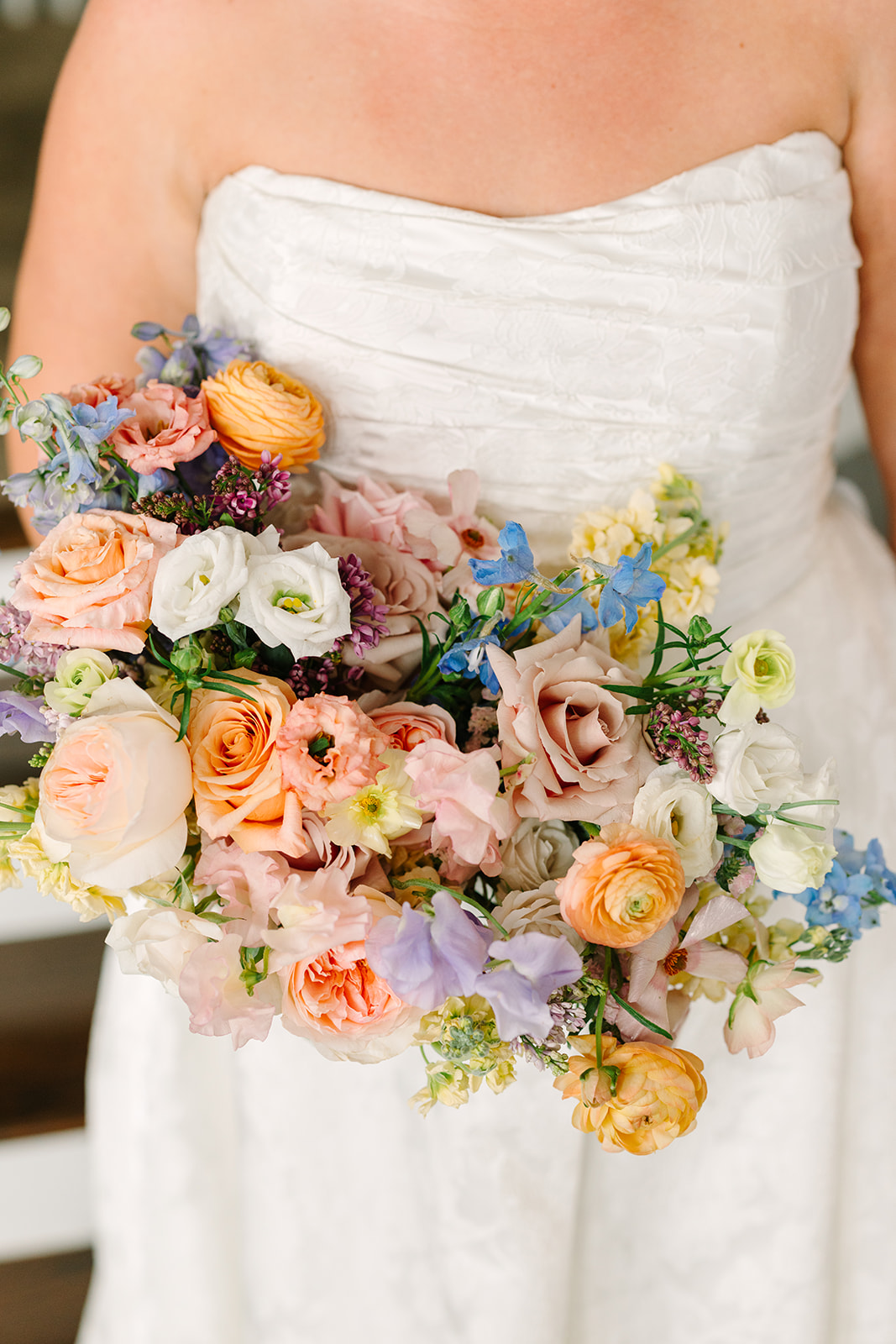 Person in a white strapless dress holding a bouquet of pastel-colored flowers, including roses and ranunculus.