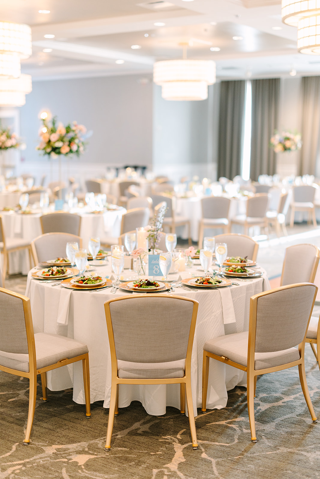 A round banquet table with elegant place settings, salads on plates, and a tall floral centerpiece; similar tables fill a bright, decorated event hall.