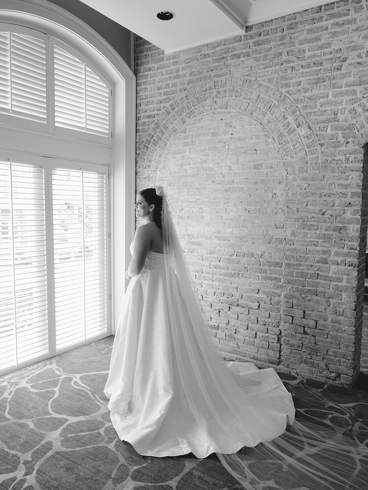 A bride in a strapless wedding gown and long veil stands indoors near large windows, facing away from the camera, with exposed brick walls in the background.