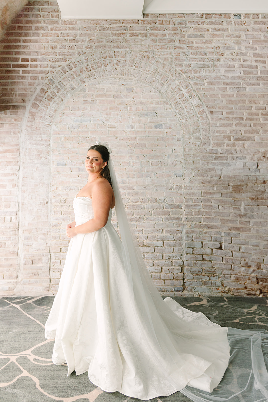A woman in a white wedding dress and veil stands smiling in front of a light-colored brick wall.