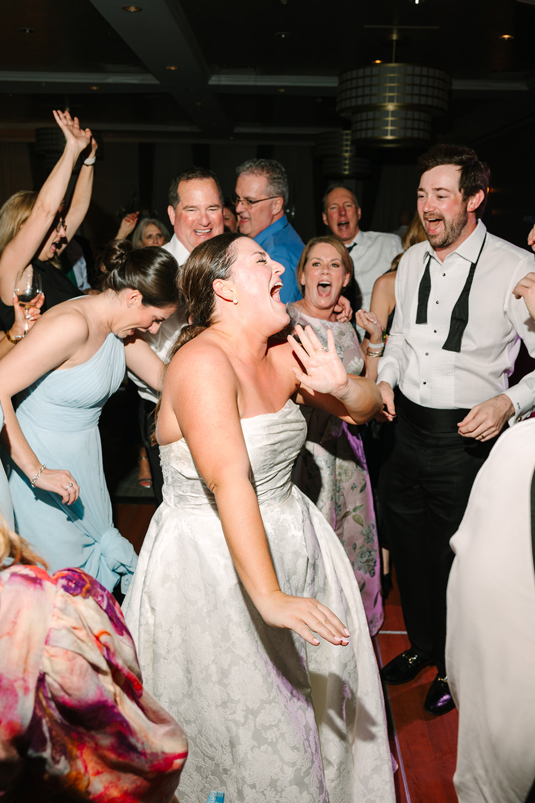 A group of people wearing colorful dresses dance on a wooden floor under modern chandeliers 
