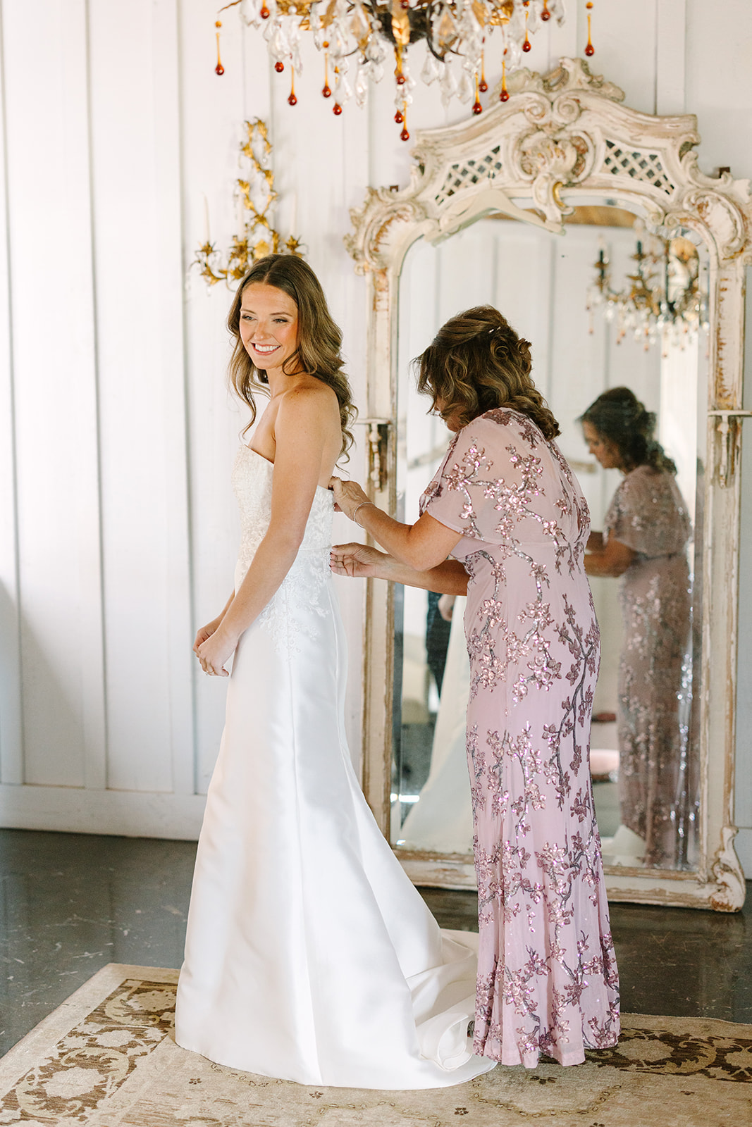 A woman in a white wedding dress is being helped with the back of her gown by another woman in a mauve dress, standing in front of a large ornate mirror.