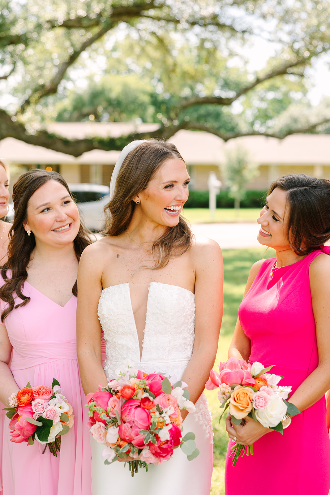 A group of women in pink and white dresses stand outdoors under large trees, holding bouquets of flowers and posing for a photo for a pink summer wedding