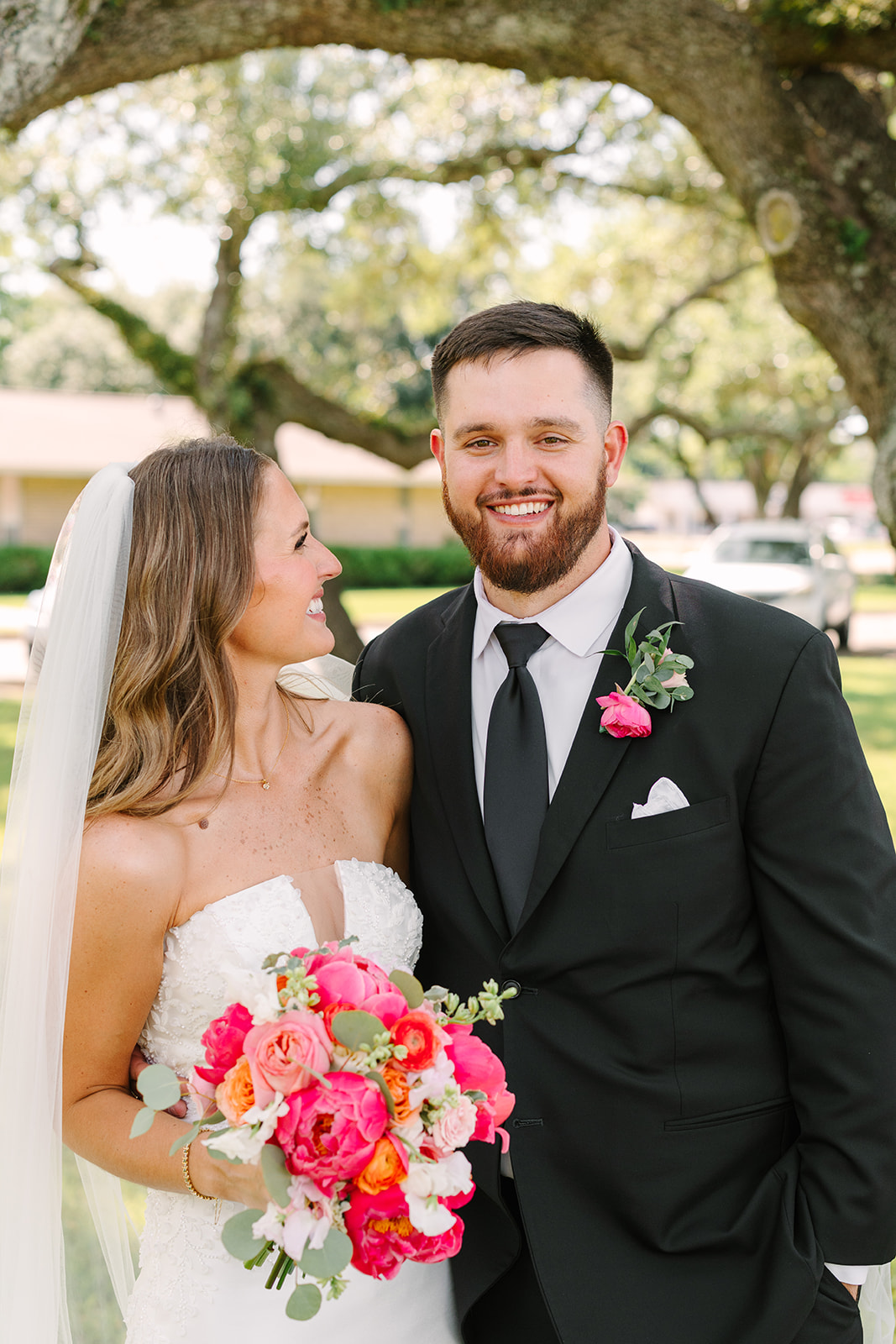 A bride and groom in wedding attire stand close together, smiling and posing for a photo under large, leafy trees in an outdoor setting for a pink summer wedding