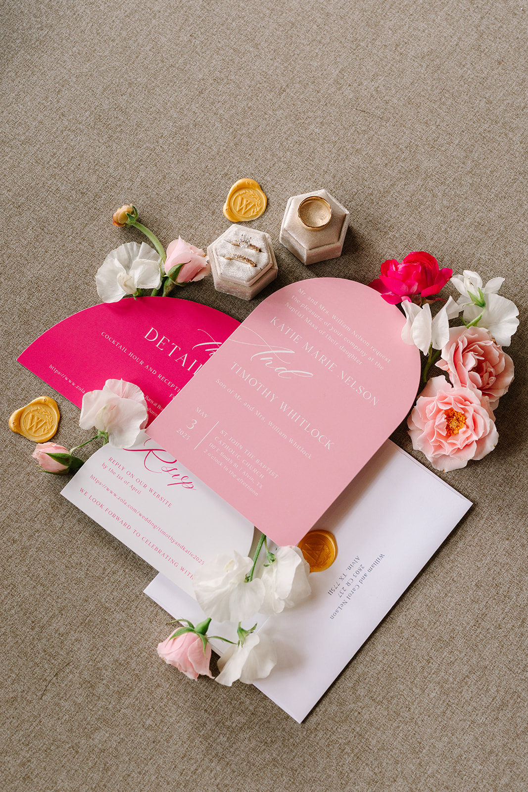 Three pink and white wedding invitations with gold wax seals and assorted flowers arranged around them on a beige surface for a pink summer wedding