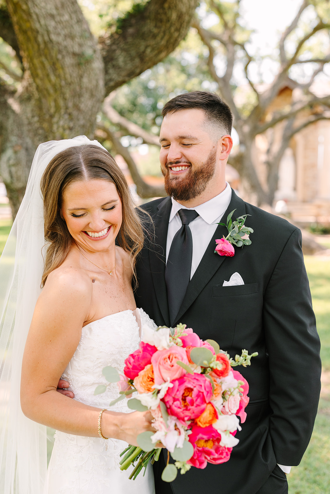 A bride and groom in wedding attire stand close together, smiling and posing for a photo under large, leafy trees in an outdoor setting for a pink summer wedding