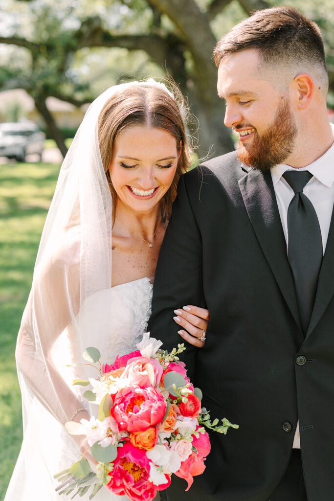 A bride and groom in wedding attire stand close together, smiling and posing for a photo under large, leafy trees in an outdoor setting for a pink summer wedding