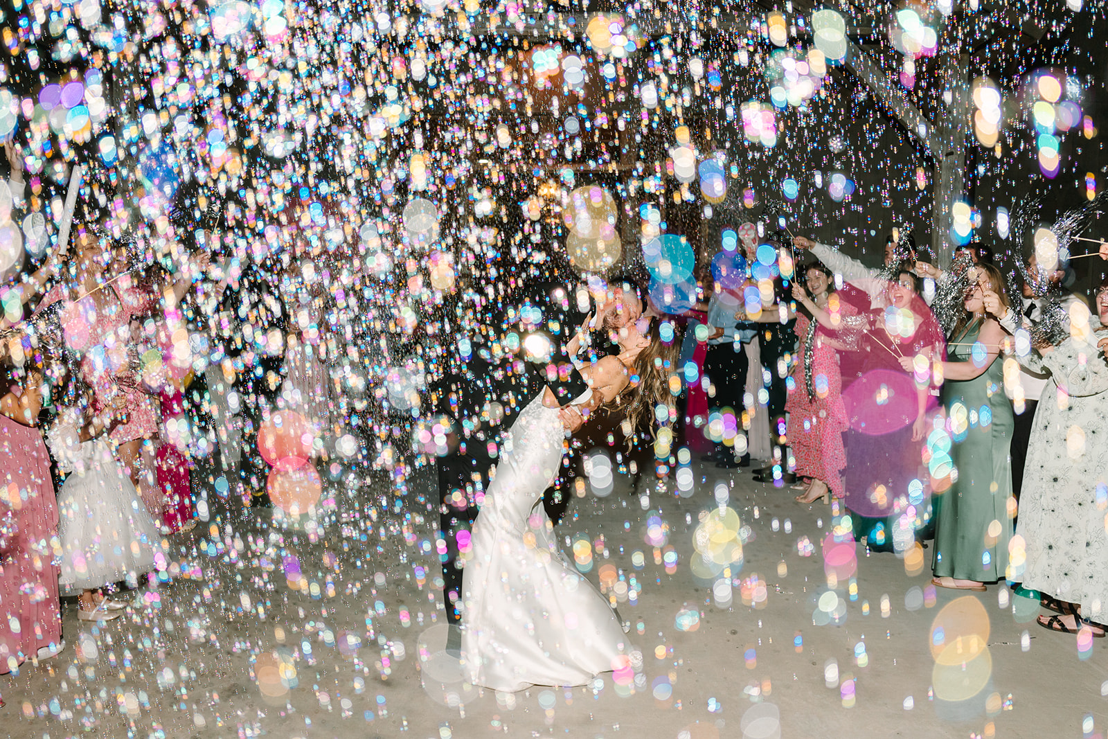 A bride and groom walk outside smiling with their hands raised as guests celebrate and blow bubbles around them.