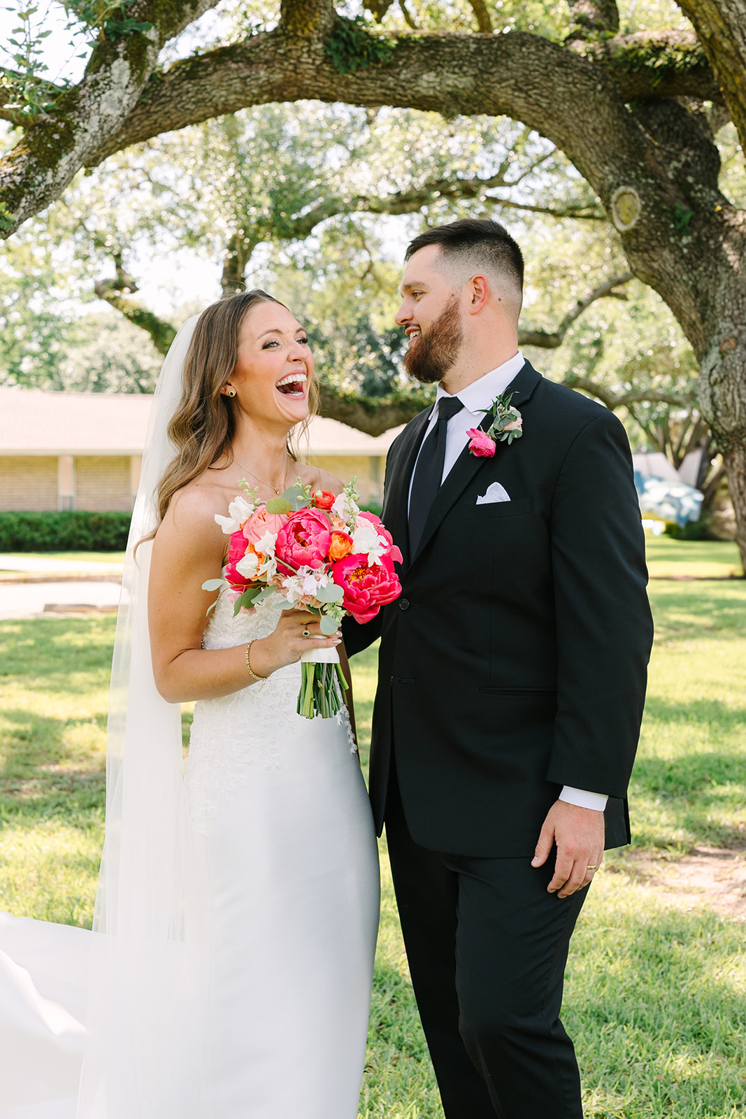 A bride and groom in wedding attire stand close together, smiling and posing for a photo under large, leafy trees in an outdoor setting for a pink summer wedding