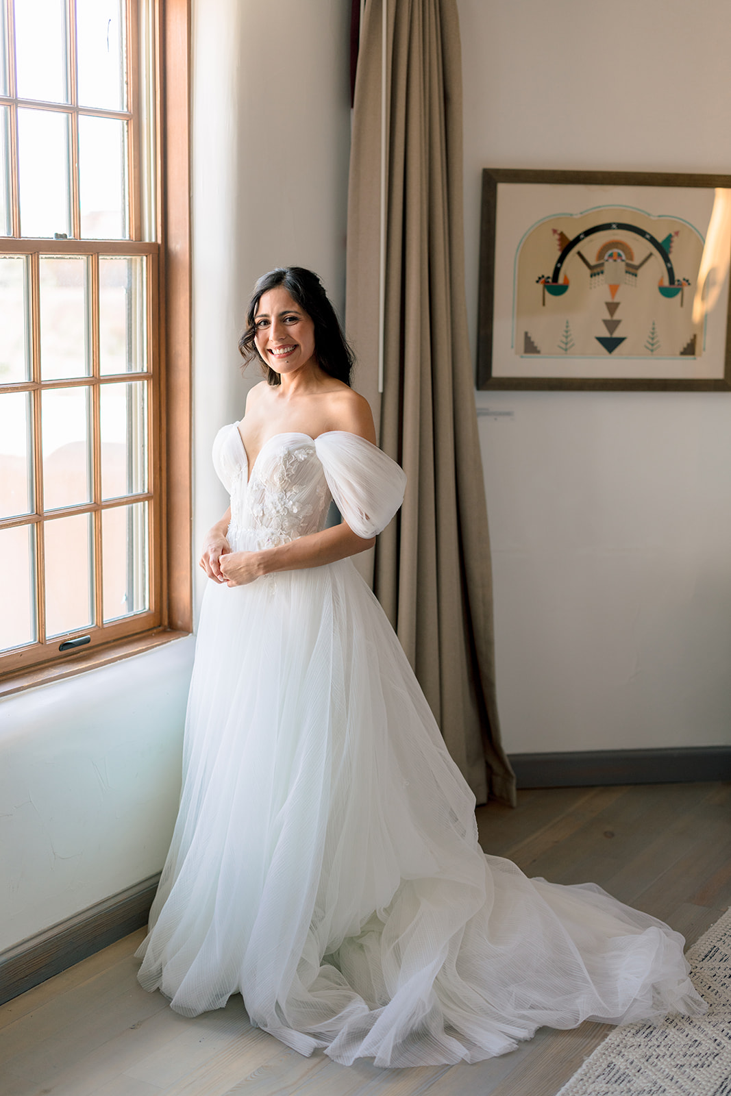 A woman in an off-the-shoulder white wedding gown stands by a window, smiling, with framed artwork and beige curtains in the background.