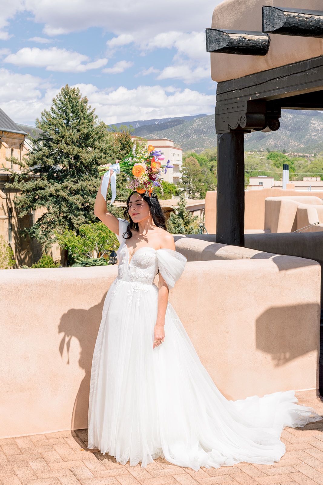 A woman with dark hair in a white off-the-shoulder wedding dress stands against a plain white background, smiling and looking slightly to the side.