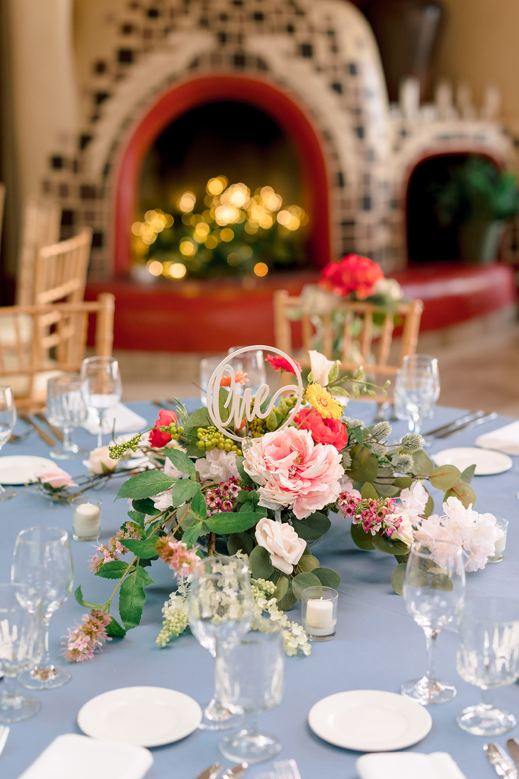 Round tables with blue tablecloths and floral centerpieces are set up in a room with a tiled fireplace and wooden chairs at La Fonda