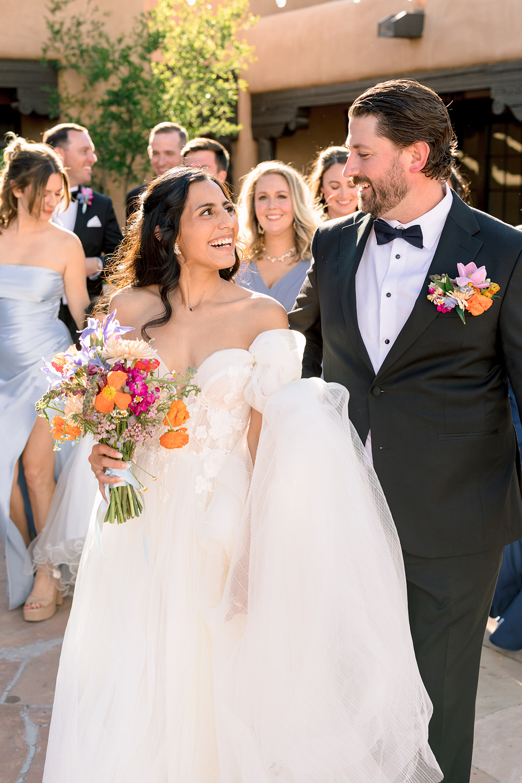 A bride and groom walk together outdoors, surrounded by their wedding party holding colorful bouquets, with a flower girl in a white dress at the front.