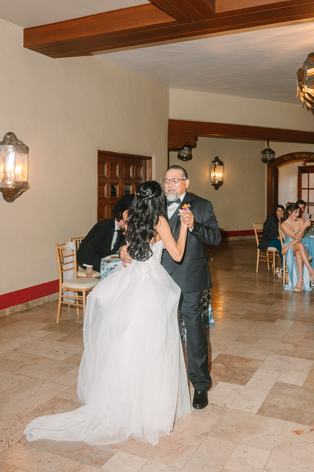 A bride and groom hold hands and enter a venue; the bride raises one arm in celebration, both are dressed formally.