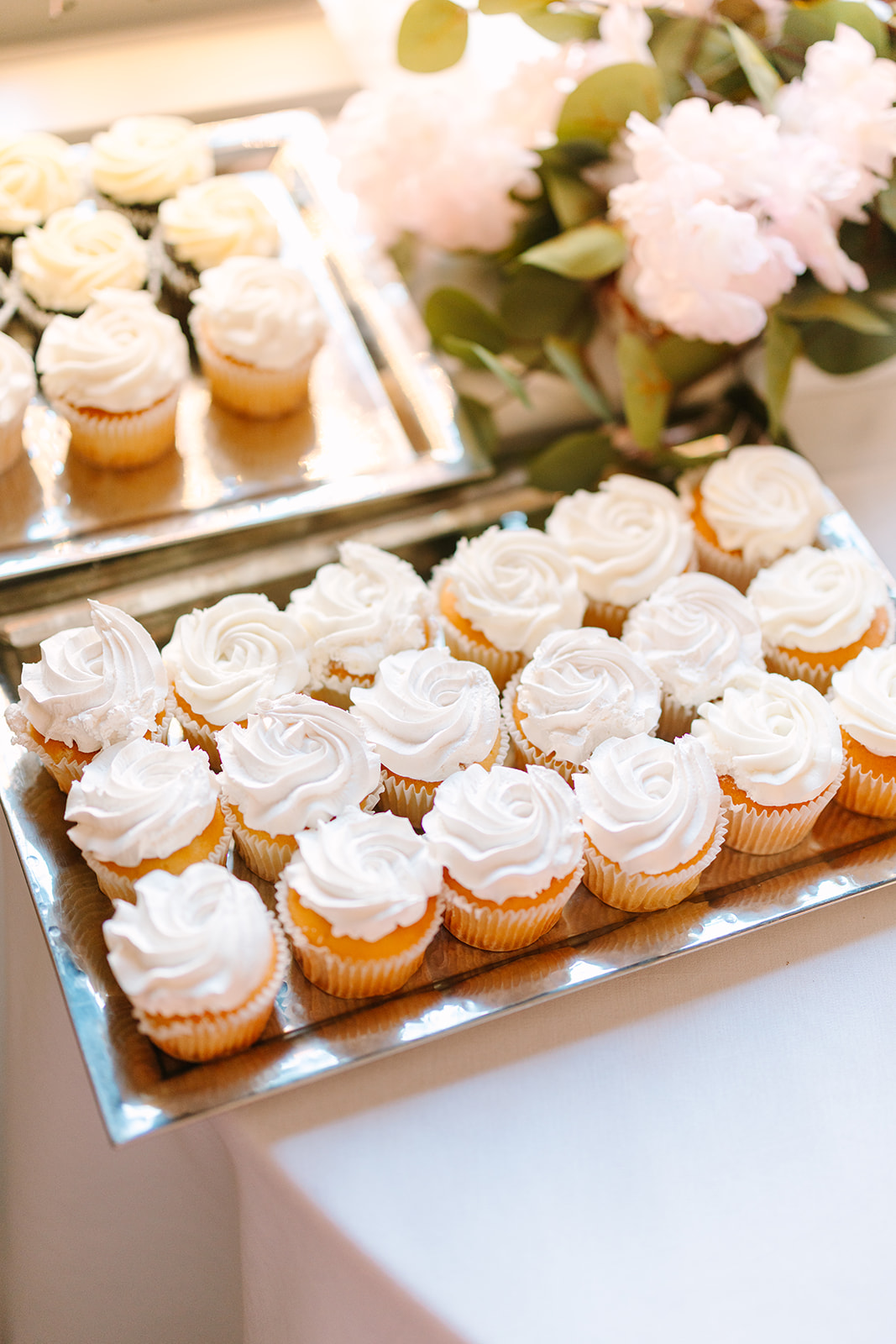 A two-tier cake with a bride and groom topper sits on a gold stand next to a tray of cupcakes with white frosting, surrounded by candles and floral decorations.