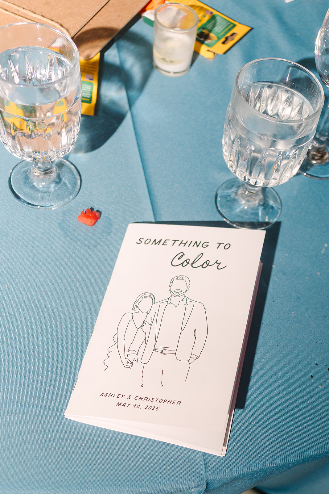 A booklet titled "Something to Color" lies on a blue tablecloth next to two crystal glasses and a small red crayon.