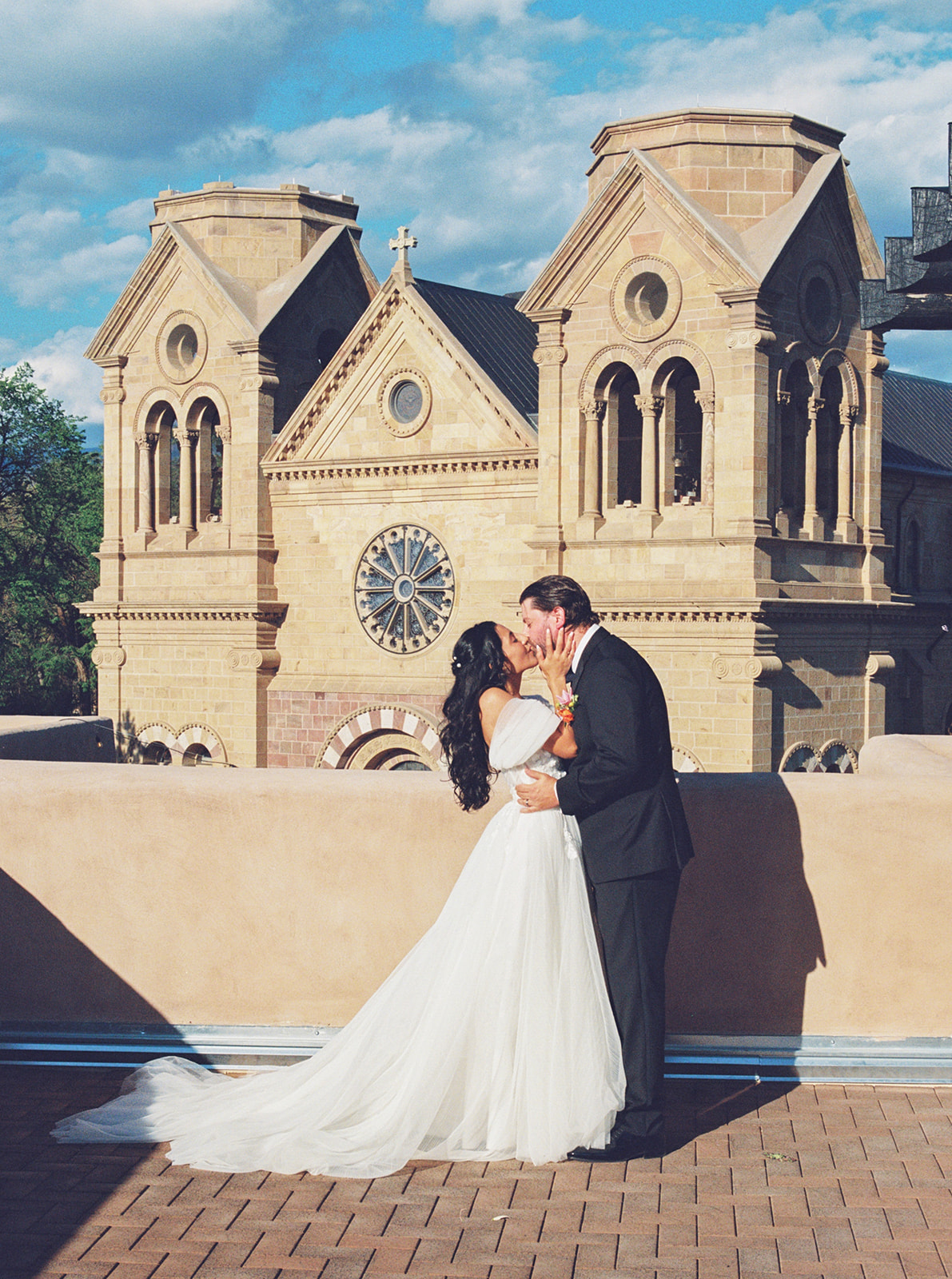 A bride and groom kiss on a terrace with a historic church building in the background under a partly cloudy sky.