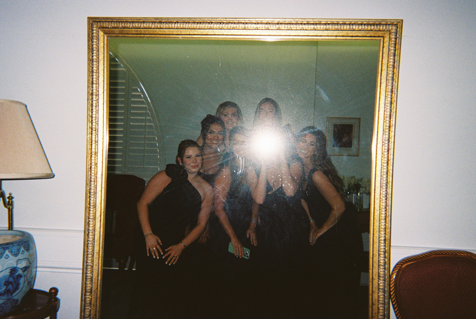 Six women in dark dresses pose together in front of a large mirror; the camera flash obscures their faces.