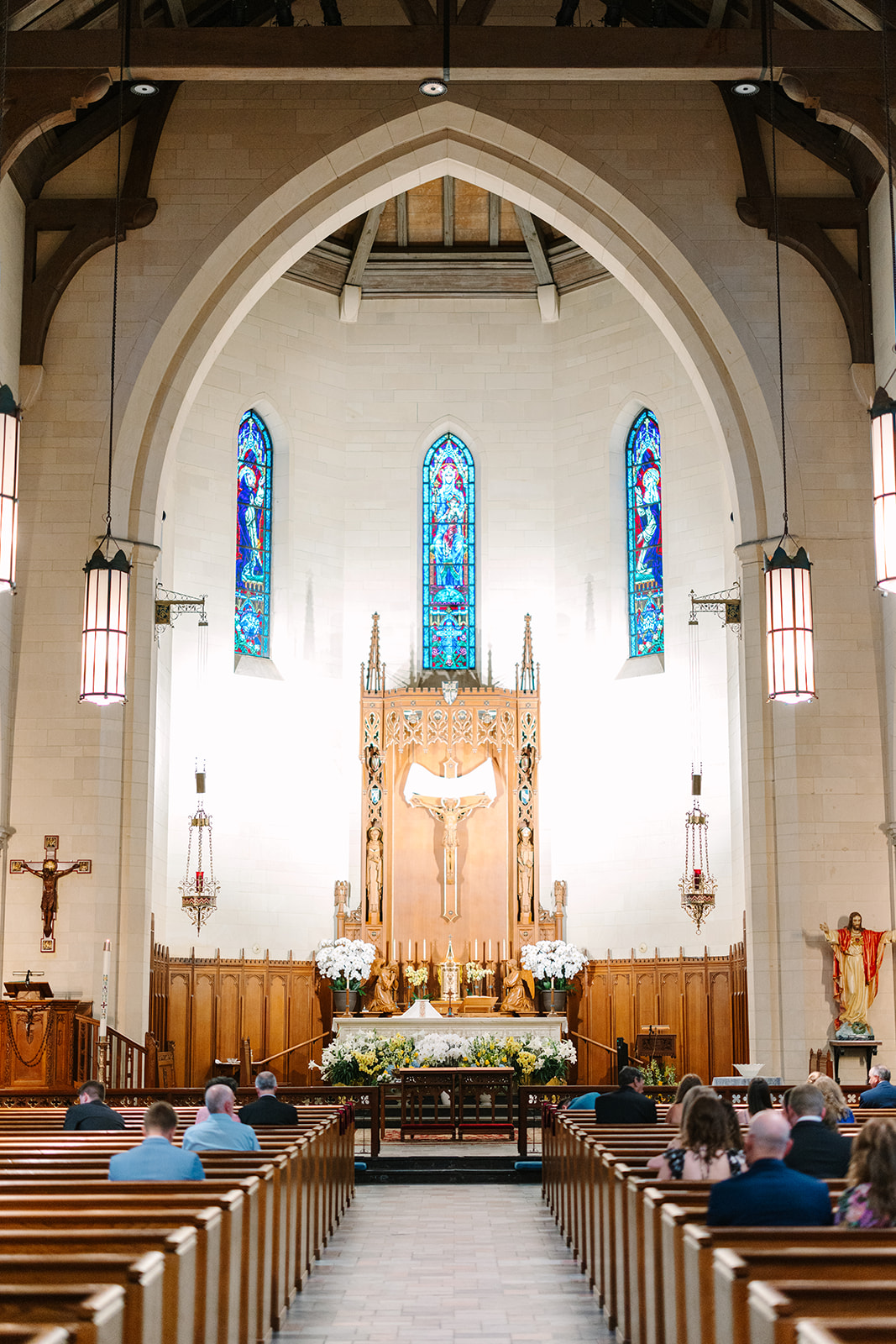 Interior of a church with stained glass windows, wooden pews, an altar decorated with flowers, and several people seated facing the front 