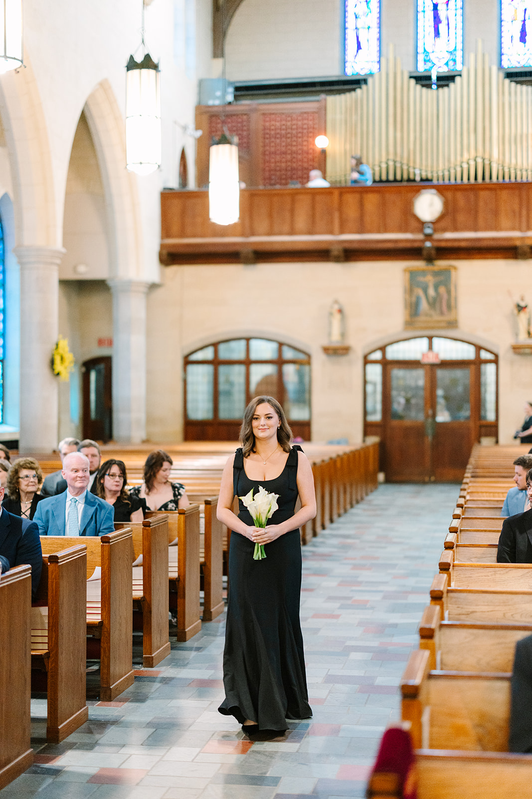 A woman in a black dress holds a bouquet of flowers while walking down the aisle of a church filled with seated guests.